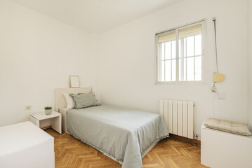 Small room with a single bed with a gray bedspread and matching cushion with an aluminum radiator, windows and aluminum bars