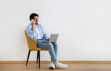 Cheerful young muslim guy in casual and glasses sits on armchair with laptop, calls by phone in room