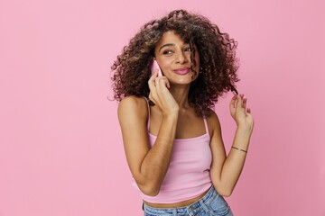 Woman with curly afro hair talking on phone in pink top and jeans on pink background, smile, happiness, copy space