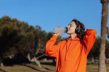Active young woman in wireless headphones drinks water from bottle after exercising outdoors at park. Female athlete in orange sweatshirt quenches thirst and cools down after training workout outside