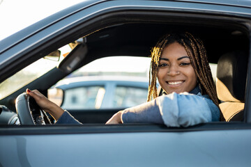 Dark-skinned young woman with braids sitting in a car looking into the camera smiling.Testing a new car, novice drivers. Travel concept of African women.