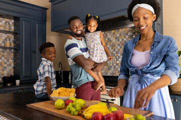 African american smiling husband and children looking at woman chopping fruits on kitchen counter