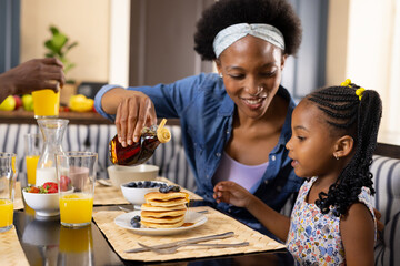 African american mother pouring honey on pancakes while having breakfast with daughter at home