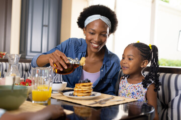 African american smiling mother pouring honey on pancakes while having breakfast with daughter