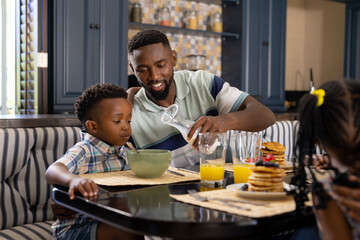 Fototapeta na wymiar African american father pouring milk in bowl for son while having breakfast at dining table