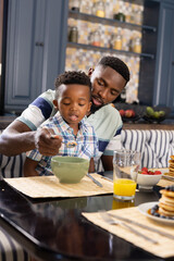 African american father feeding breakfast cereals to son at dining table at home, copy space