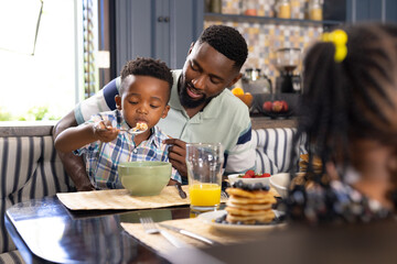 African american father looking at son eating breakfast cereals at dining table at home, copy space