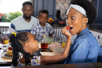 African american daughter feeding strawberry to mother while having breakfast with family at home