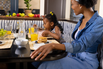 African american mother looking at daughter eating pancakes and juice at dining table at home