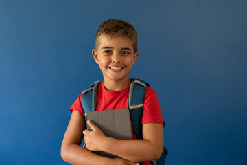 Caucasian school boy with backpack and digital tablet standing on blue background, copy space