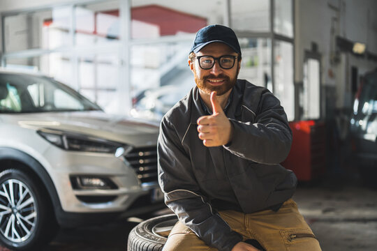 smiling Caucasian auto mechanic with glasses sitting on wheels and showing thumbs up at workshop, medium shot. High quality photo