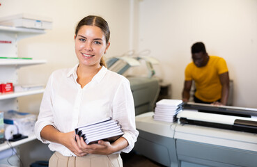 Cheerful young woman in white shirt smiling at the camera and holding new planners in the printing...