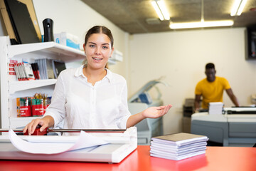 Cheerful young woman in white shirt smiling at the camera and using paper cutter in the printing house