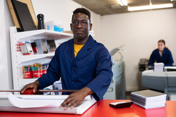 Serious middle-aged African American male in a blue uniform using paper cutter in the printer house