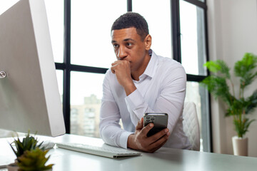 puzzled african american man in business clothes sits in the office at the table holds smartphone