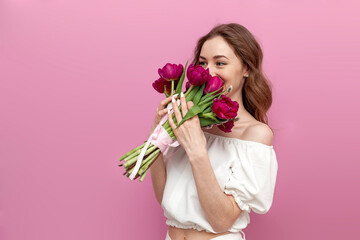 cute young woman in festive outfit holds bouquet of pink tulips and smells them on pink isolated...