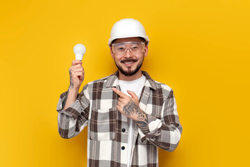 male asian electrician in hard hat and glasses holds light bulb and points at it with finger on...