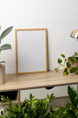 Vertical of empty wooden frame with copy space, lamp and plants in pots on table against white wall