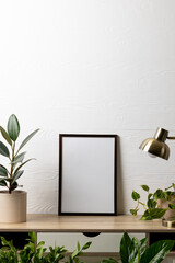 Vertical of black empty frame with copy space, lamp and plants in pots on table against white wall