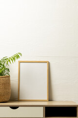 Vertical of empty wooden frame with copy space and plant in pot on desk against white wall