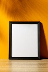 Vertical of black empty frame with copy space on table against yellow wall and shadow of palm leaves