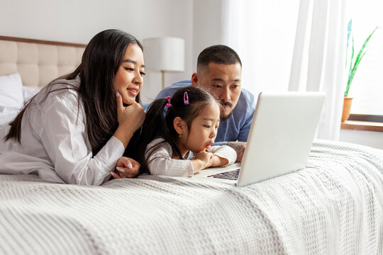 Asian Family Use Laptop At Home On Bed, Korean Dad Mom And Little Daughter Look At Computer Screen And Study Online