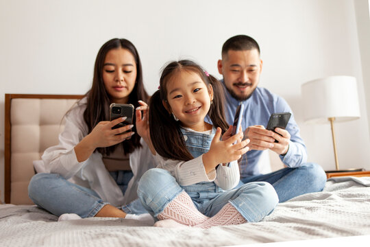little asian girl with mom and dad use smart phone, korean happy young family with mobile gadgets sit on bed at home