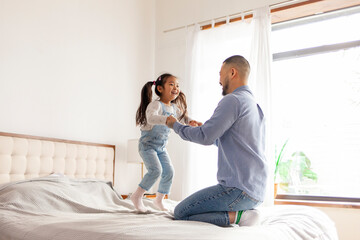 small active Asian girl plays with her dad at home, energetic child jumps and dances on the bed...