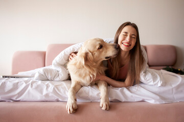 girl lies in bed and hugs golden retriever dog, the dog licks the owner, the woman loves pets - obrazy, fototapety, plakaty