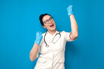woman doctor in medical gown and glasses rejoice in victory on blue isolated background, girl nurse...