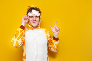 young joyful guy in funny baby giraffe pajamas and sleep mask advertises copy space on yellow background