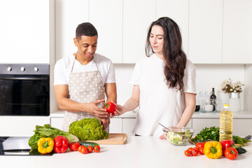 multiracial young couple preparing veggie vegetable and greens salad in white modern kitchen