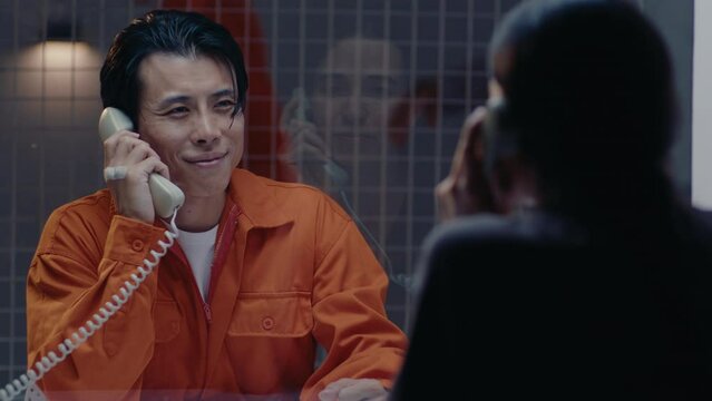 Asian male prisoner in orange jail uniform smiling and speaking on the phone with a loving wife in the visiting room. Over the shoulder, zoom shot