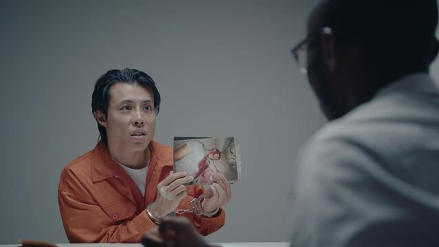 Aggressive Asian murderer in orange jumpsuit and handcuffs showing crime scene photos to detective, telling how he killed a victim, giving confession during interrogation. Over the shoulder shot