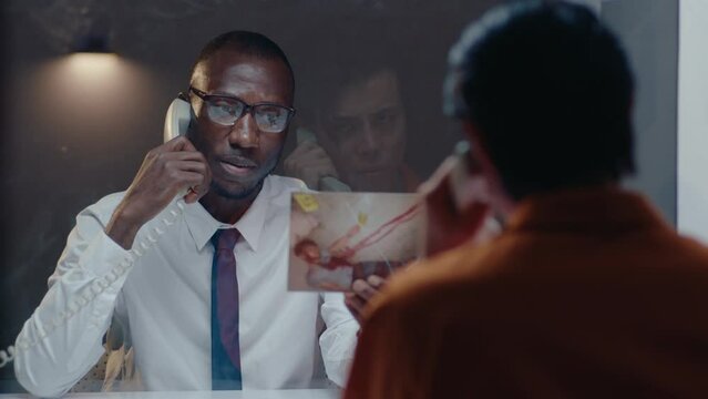 African American detective showing crime scene photo through the glass wall, speaking with prisoner on the phone in the visiting room, questioning him about murder