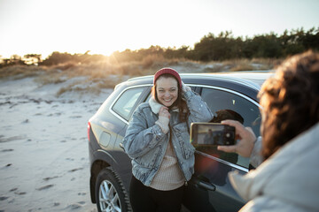Young adult man taking a picture of a young adult woman next to a car on a beach during winter