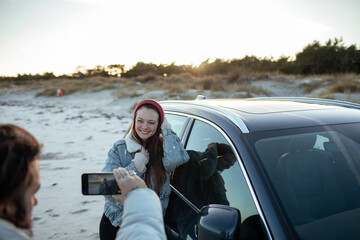 Young adult man taking a picture of a young adult woman next to a car on a beach during winter