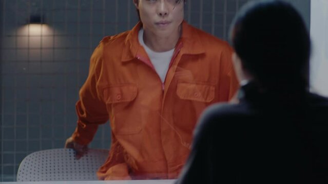 Asian prisoner in orange jumpsuit entering visiting room, sitting on chair behind the glass, talking on the phone with loving wife