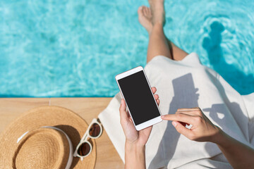 Asian woman sitting by the pool while using mobile phone. Young traveler female working on cellphone during her summer vacation trip. Technology and lifestyle concept. Copy space, top view
