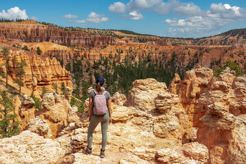 Woman with backpack looking at scenic aerial view of hoodoo sandstone rock formations on Peekaboo hiking trail in Bryce Canyon National Park, Utah, USA. Natural amphitheatre on sunny summer day