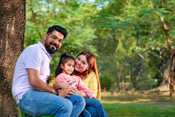 Naklejka na ściany i meble Happy Young indian parents with their cute little daughter sitting at park or garden.