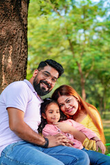 Happy Young indian parents with their cute little daughter sitting at park or garden.