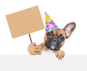 French bulldog puppy wearing  party cap looks above empty white banner and shows empty placard. isolated on white background