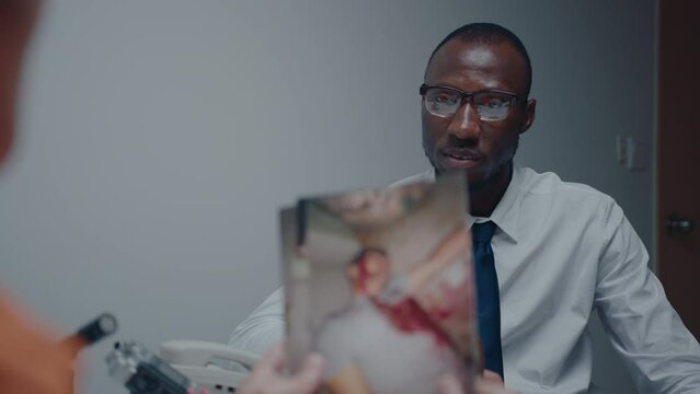 African American detective questioning killer as he watching crime scene pictures during interrogation. Over the shoulder shot
