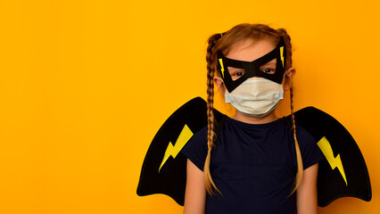 Child in a Superhero costume in a carnival and medical mask with wings behind his back on an orange background. Little cute strong girl with pigtails bravely fights disease and stands on the defense
