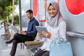 Smiling Asian woman in hijab holding digital tablet sitting at bus stop with a background of men sitting waiting for the bus together