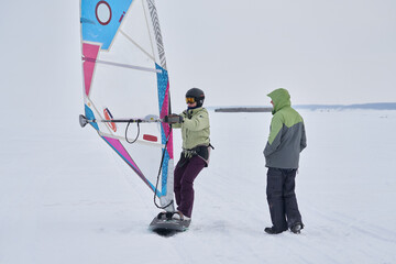 A man, a snowsurfer, helps a woman, his wife, start riding a sailboard. A middle-aged man and woman go snowsurfing on a cloudy winter day.