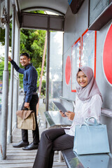 Asian man stands and woman in hijab sits smiling at camera holding tablet at bus stop