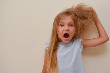 A little pretty girl with an open mouth in horror holds with her hand half of her hair, which is very tangled, the other half of her hair is combed and lies silky. Child shocked by a mess on the head