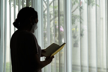 Silhouette of a woman who is standing by the window reading a book - obrazy, fototapety, plakaty